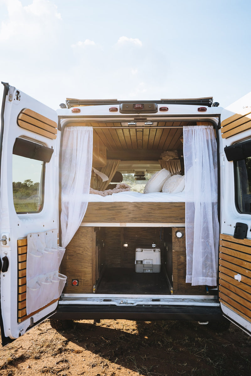 Inside an RV rental camper van: open rear doors reveal a cozy bed, sheer curtains, and a portable toilet on a pull-out tray. — photo 12