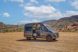 This gray custom camper van rv rental sits in a scenic mountain landscape. Its open door reveals a modern interior, ready for adventure.