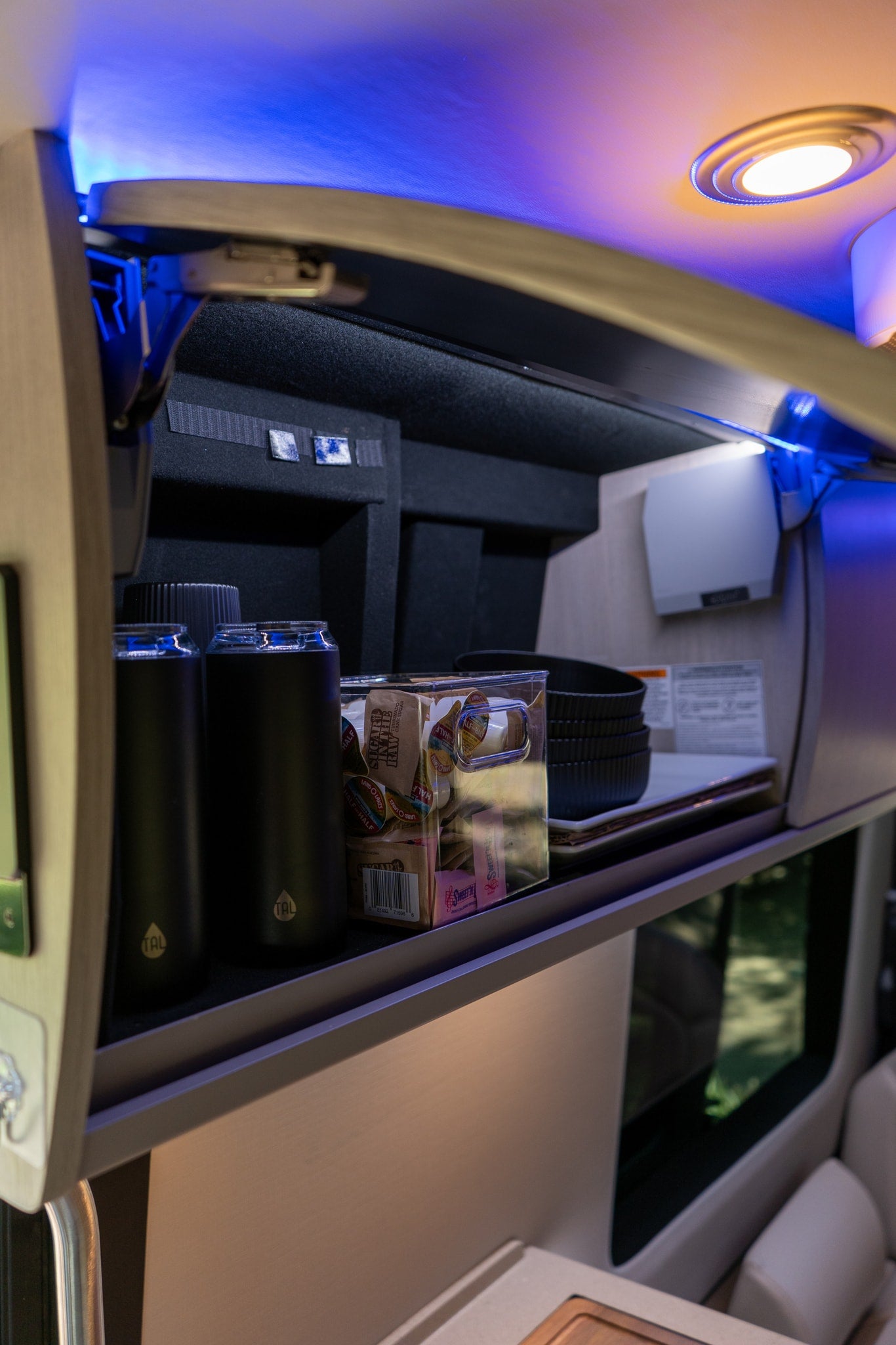 RV rental overhead cabinet, lit by blue LEDs, holds TAL water bottles, sugar/creamer packets, and bowls for organized travel. — photo 15