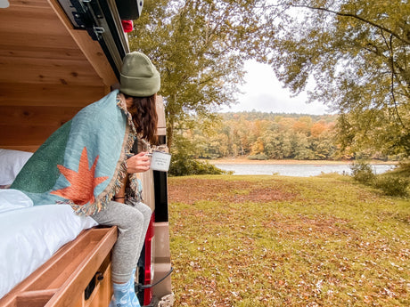 Cozy fall morning view from an RV rental, person wrapped in blanket enjoying coffee by the river.