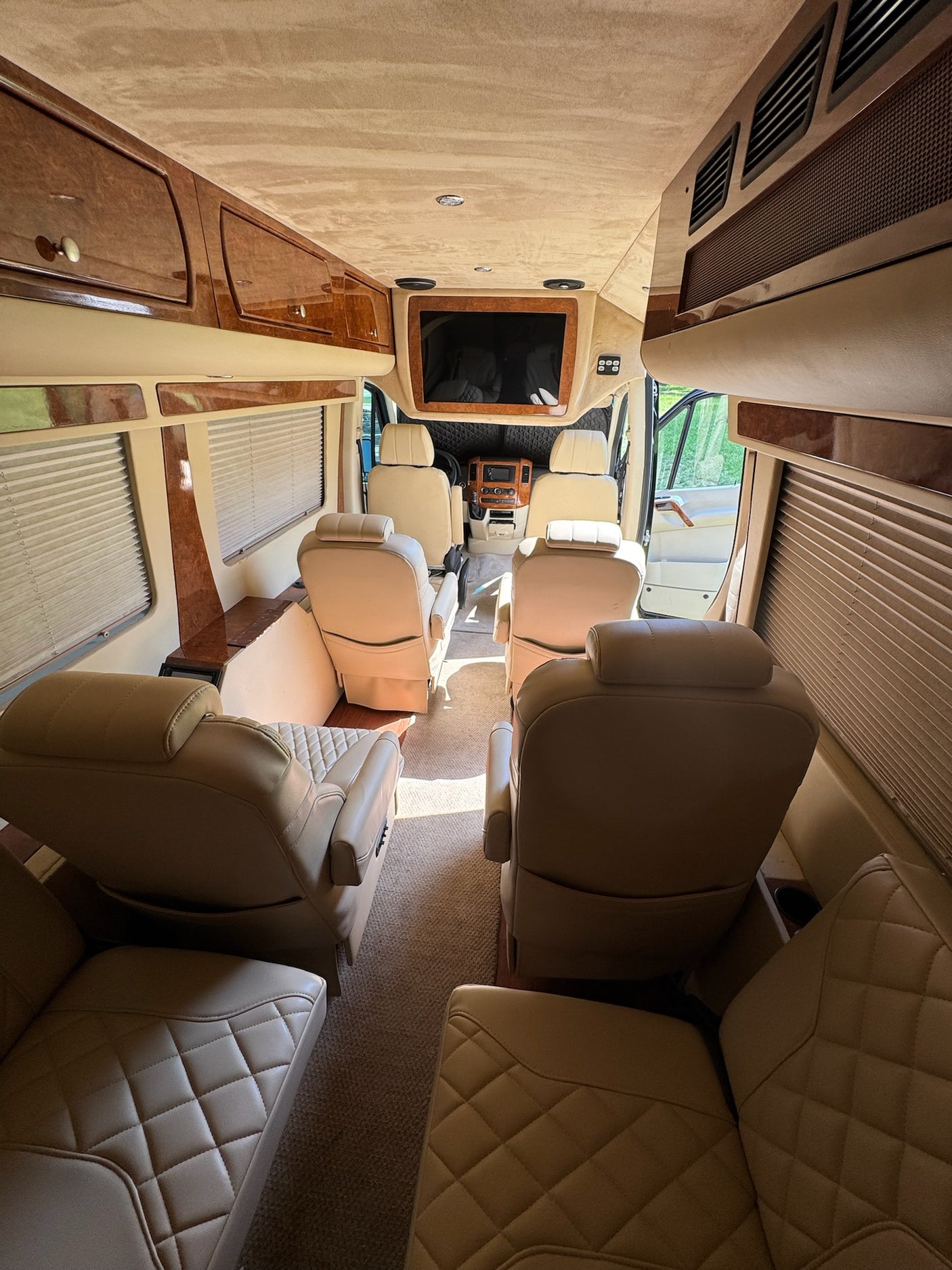 Step inside this luxurious RV rental: a spacious interior with multiple rows of comfortable, quilted tan leather seats, rich wood trim, and a built-in TV for premium travel comfort.