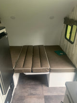 RV rental: Compact camper interior with brown upholstered bed/seating, compact refrigerator, and window view.