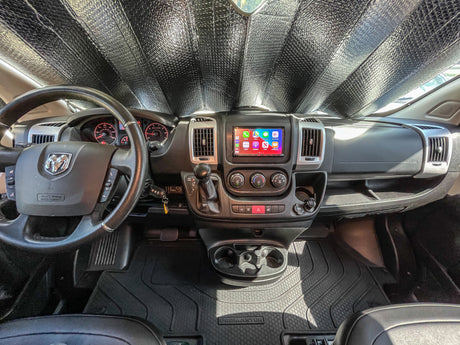 Inside a Ram ProMaster rv rental van: dashboard view with a sunshade, steering wheel, infotainment, and floor mats.
