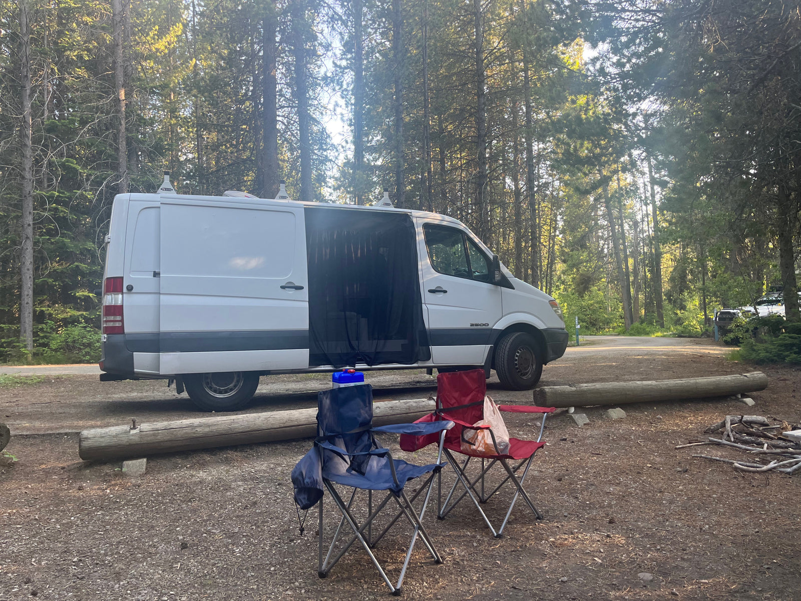 Forest campsite scene: white RV rental van with mesh screen, blue & red camping chairs, & firewood. Perfect for glamping.