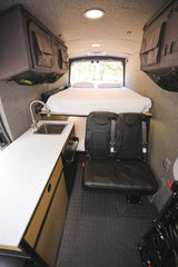 An RV rental's cozy camper van interior: a comfortable bed, compact kitchen with sink, and two passenger seats.