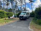 Gray rv rental van parked at a forest campsite. A person cooks at a picnic table, with a smoking fire pit nearby.
