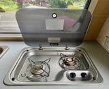 Inside an RV rental: a compact stainless steel Dometic two-burner gas stove with its glass lid open, beside a small sink.