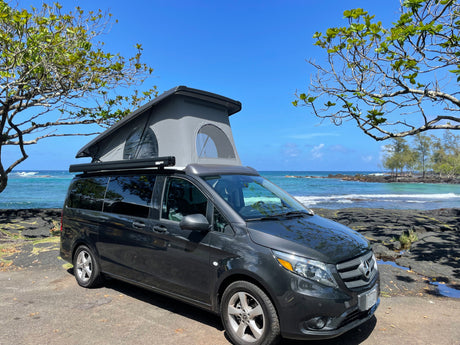 RV rental: Dark grey Mercedes-Benz campervan with pop-up roof, parked by a blue ocean with rocky shores under clear skies.