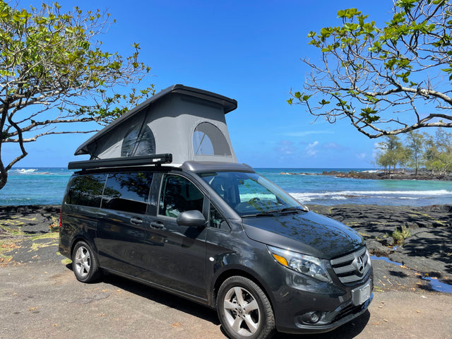 RV rental: Dark grey Mercedes-Benz campervan with pop-up roof, parked by a blue ocean with rocky shores under clear skies.