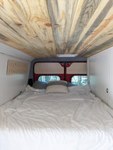 Comfortable bed inside a modern rv rental camper van, featuring a unique wooden ceiling and scenic rear windows.
