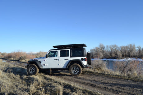 White Jeep Wrangler with rooftop tent parked off-road by a river, ideal for an outdoor rv rental adventure.