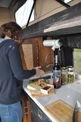 A person in an RV rental prepares snacks with cheese, wine, and olives, showcasing the compact kitchen setup.