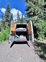 RV rental van's back open, showcasing a comfy bed, storage, and wooden interior amidst tall trees and blue sky.