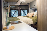 Inside a modern RV rental: a cozy bed with olive green bedding, pillows, and a kitchenette area in a spacious camper van.
