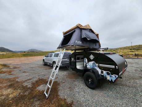 RV rental setup: Silver truck, black teardrop camper with SkyeDen rooftop tent, and ladder on gravel, ready for adventure.