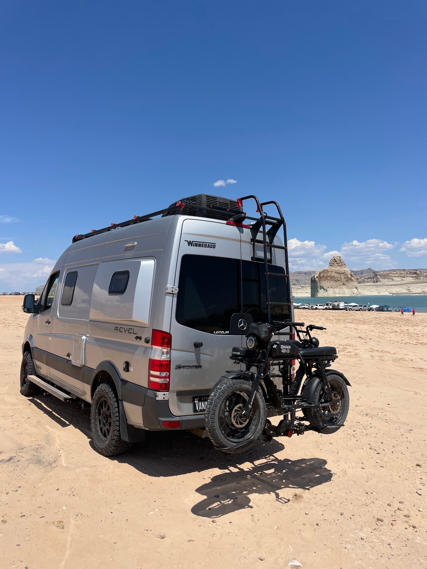 Silver Winnebago Revel RV rental van & e-bikes on a sandy lake shore under blue skies, ready for adventure. — photo 12