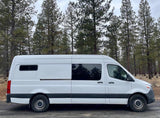 Side view of a white camper van, perfect for rv rental, parked on a dirt road by pine trees.