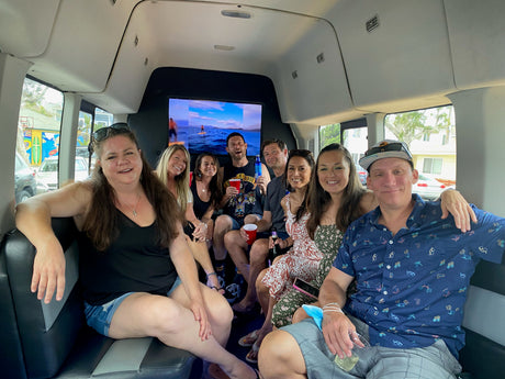 Eight friends enjoying their RV rental trip, smiling, laughing, and holding drinks inside a spacious vehicle with a TV.