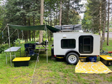 Cozy teardrop trailer RV rental setup in a wooded campsite, complete with an awning, picnic table, and outdoor rug.