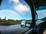 A vibrant double rainbow arches over a wet road and green trees, seen from an RV rental.