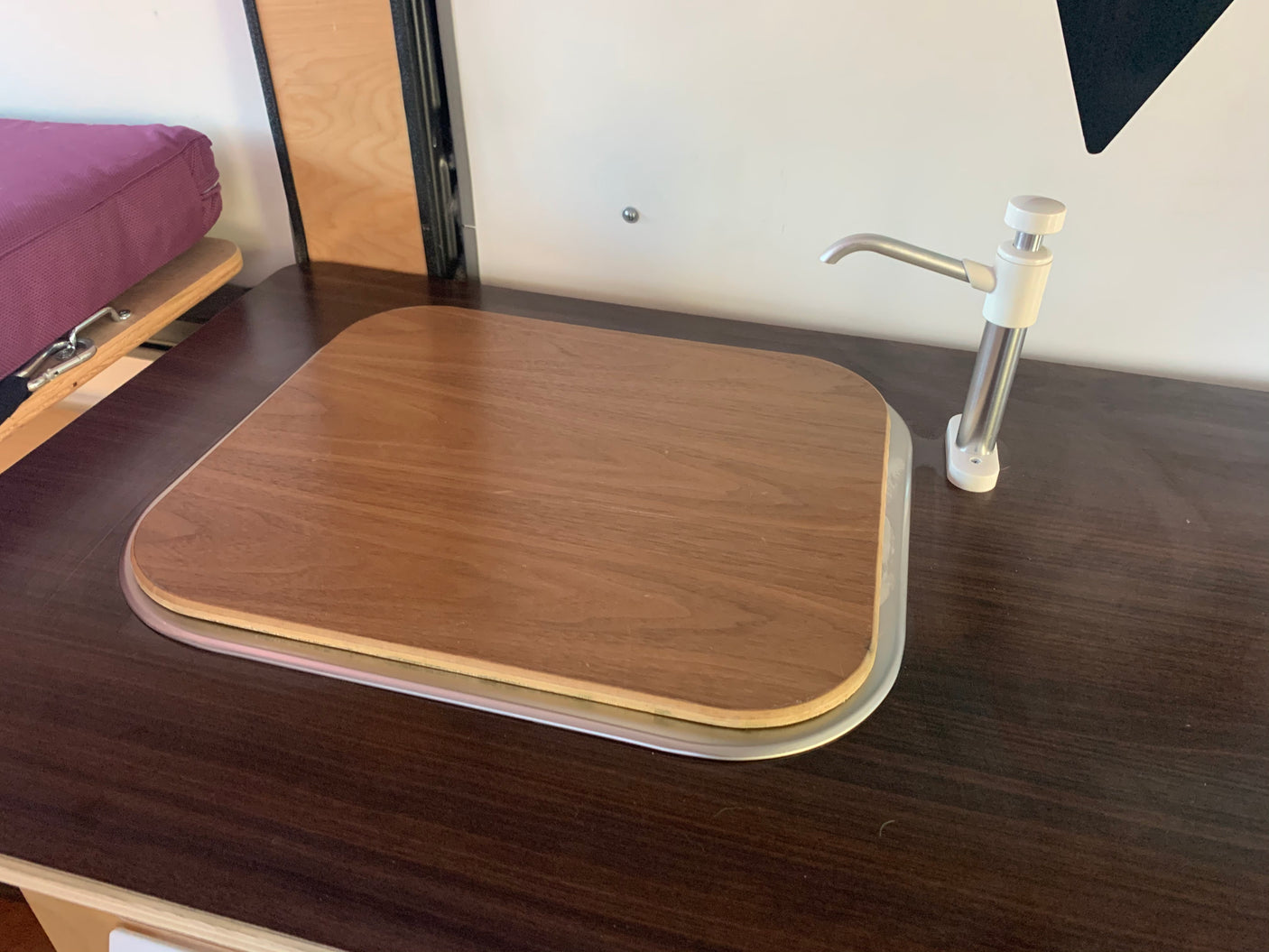 RV rental kitchenette: dark counter with a wooden sink cover and hand pump faucet. Compact and functional design.