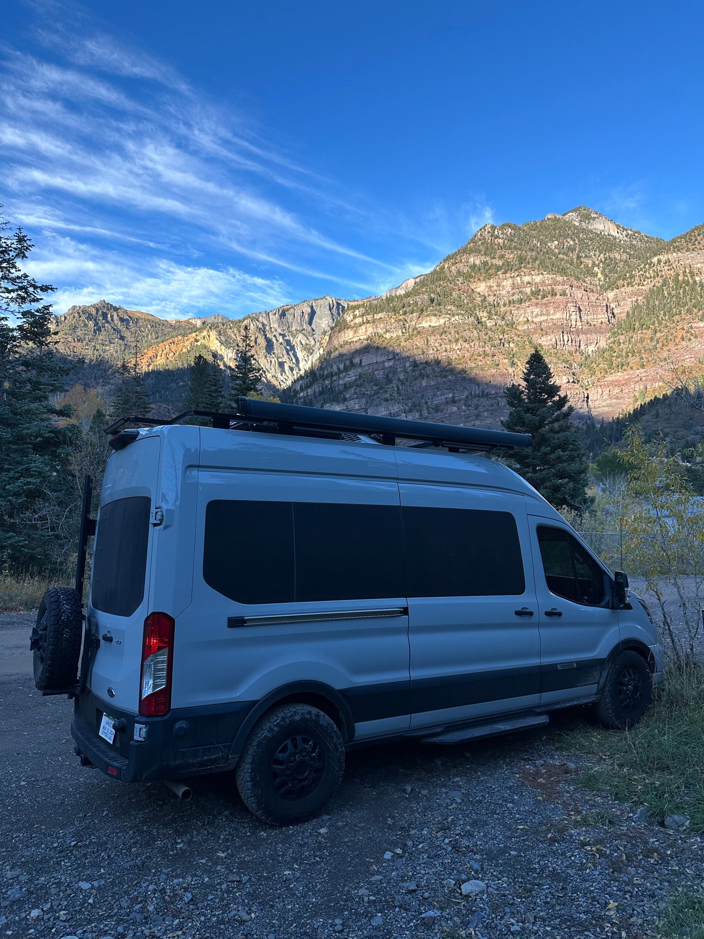 A grey camper van (rv rental) is parked on a gravel path, backed by majestic mountains and a blue sky, perfect for an outdoor adventure.