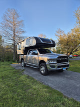 Graner rv rental: A grey Ram truck with a Palomino truck camper, parked on a paved spot amidst green grass & trees.
