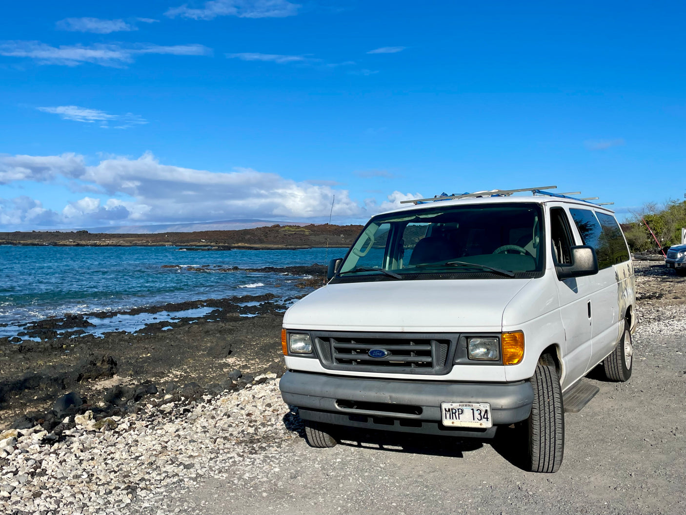 White Ford van, an RV rental, parked by Hawaii's rocky coast and blue ocean under a clear sky.