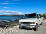 White Ford van, an RV rental, parked by Hawaii's rocky coast and blue ocean under a clear sky.