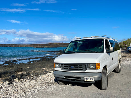 White Ford van, an RV rental, parked by Hawaii's rocky coast and blue ocean under a clear sky.