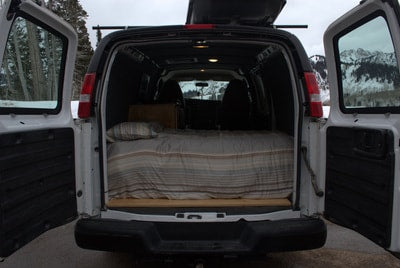 Open rear doors of a white camper van reveal a cozy bed inside, set against snowy mountains. Your next RV rental adventure awaits! — photo 13