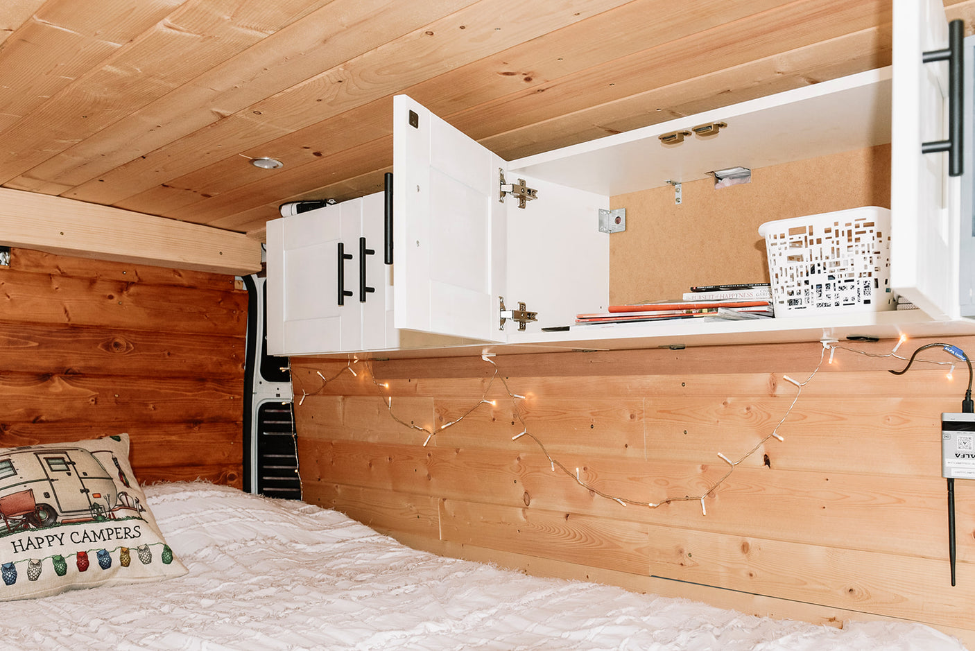 Inside an RV rental: a comfy bed with a 'Happy Campers' pillow, wooden walls, and accessible overhead cabinets.