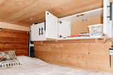 Inside an RV rental: a comfy bed with a 'Happy Campers' pillow, wooden walls, and accessible overhead cabinets.