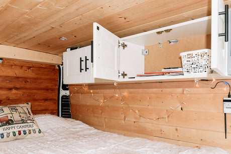 Inside an RV rental: a comfy bed with a 'Happy Campers' pillow, wooden walls, and accessible overhead cabinets.