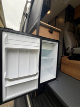 RV rental camper van interior shows an open refrigerator with shelves, along with wooden cabinetry and a passenger seat.