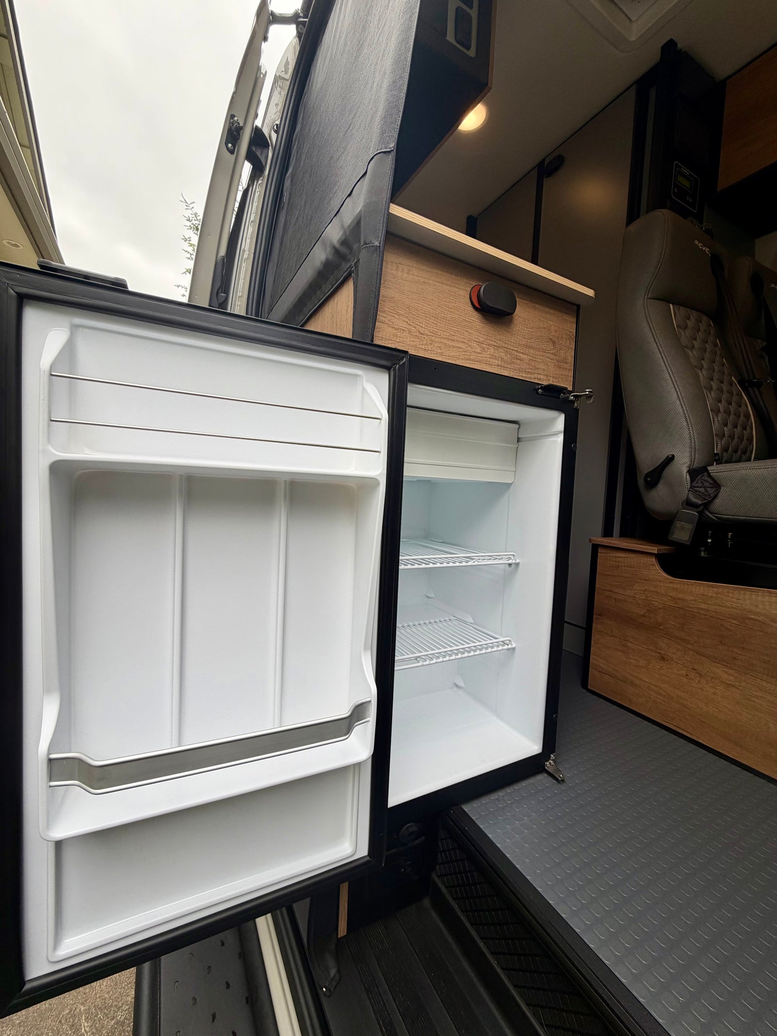 RV rental camper van interior shows an open refrigerator with shelves, along with wooden cabinetry and a passenger seat.