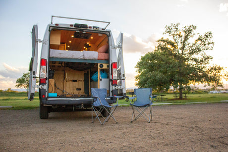 Cozy RV rental camper van with open back, revealing bed inside; two chairs are set out for camping relaxation.