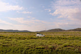A white camper van, resembling an RV rental, is centered in a wide, verdant field with distant hills under a partly cloudy sky.