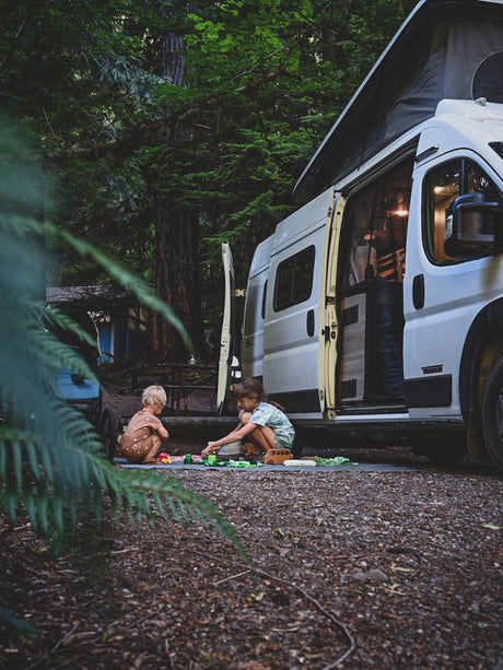Young children play outside their parked white **RV rental** at a shaded forest campsite, perfect for family adventures.