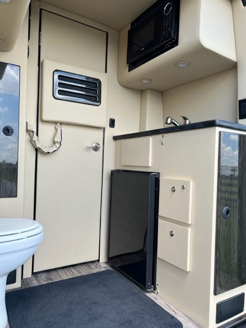 RV rental interior featuring a beige kitchen with microwave, sink, mini-fridge, and a separate bathroom door with toilet visible.