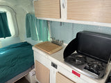 Cozy RV rental interior with a compact kitchen featuring a stove, sink, and backsplash, plus a comfortable bed.