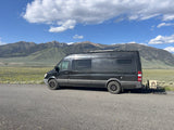 Scenic RV rental: a dark grey camper van parked on a road, overlooking mountains and a green valley under blue sky.