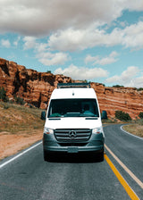 An RV rental, a white Sprinter van, on a scenic road through red rock cliffs under a cloudy sky. Ready for adventure.