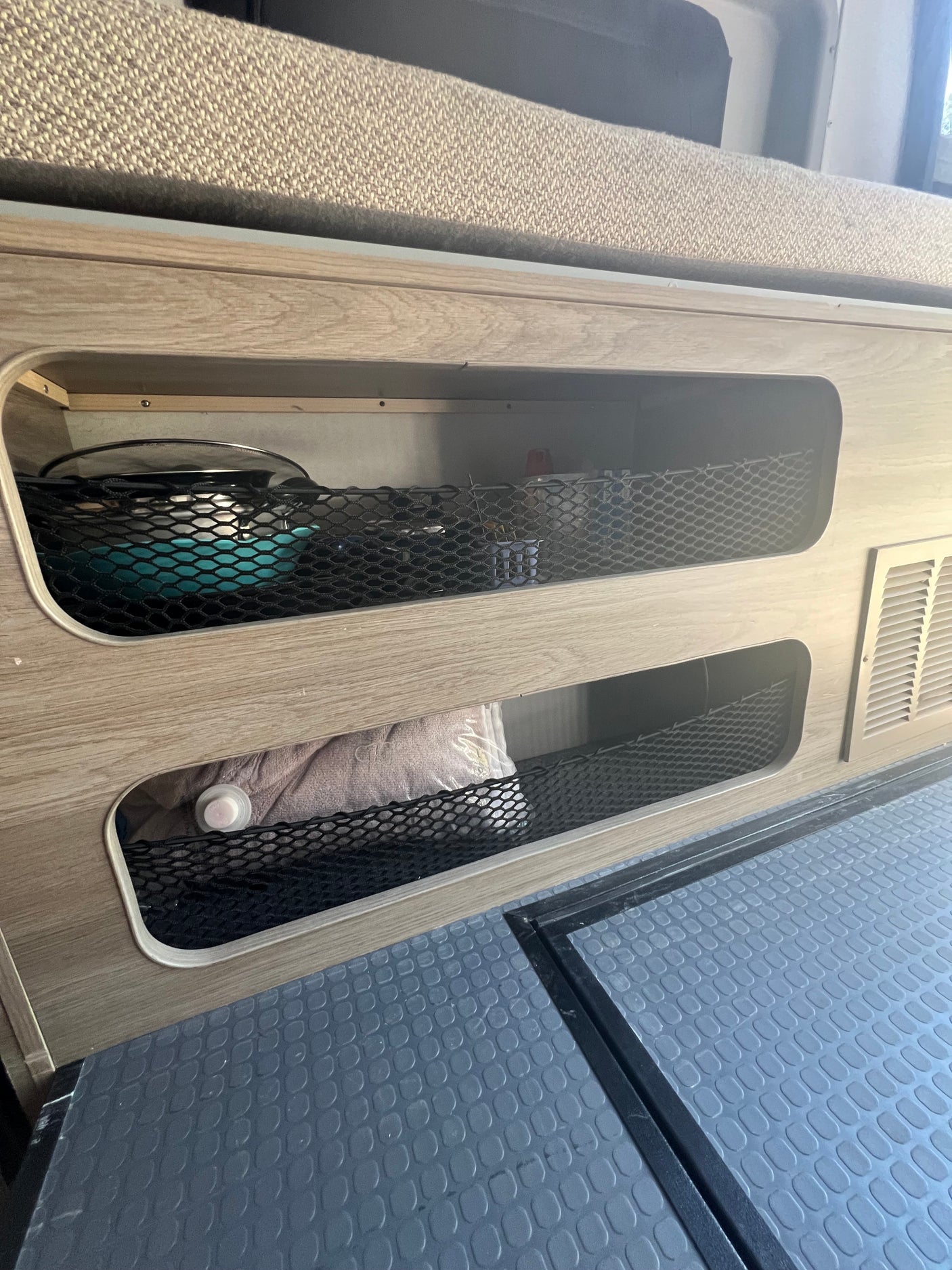 RV rental interior: Built-in wood storage with mesh nets holds kitchenware & linens. Textured floor and vent visible.