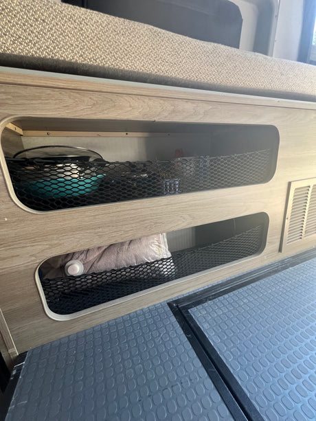 RV rental interior: Built-in wood storage with mesh nets holds kitchenware & linens. Textured floor and vent visible.