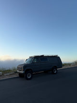 Dark grey lifted Ford Econoline camper van, ideal for your next RV rental adventure, parked by a misty hillside under blue sky.