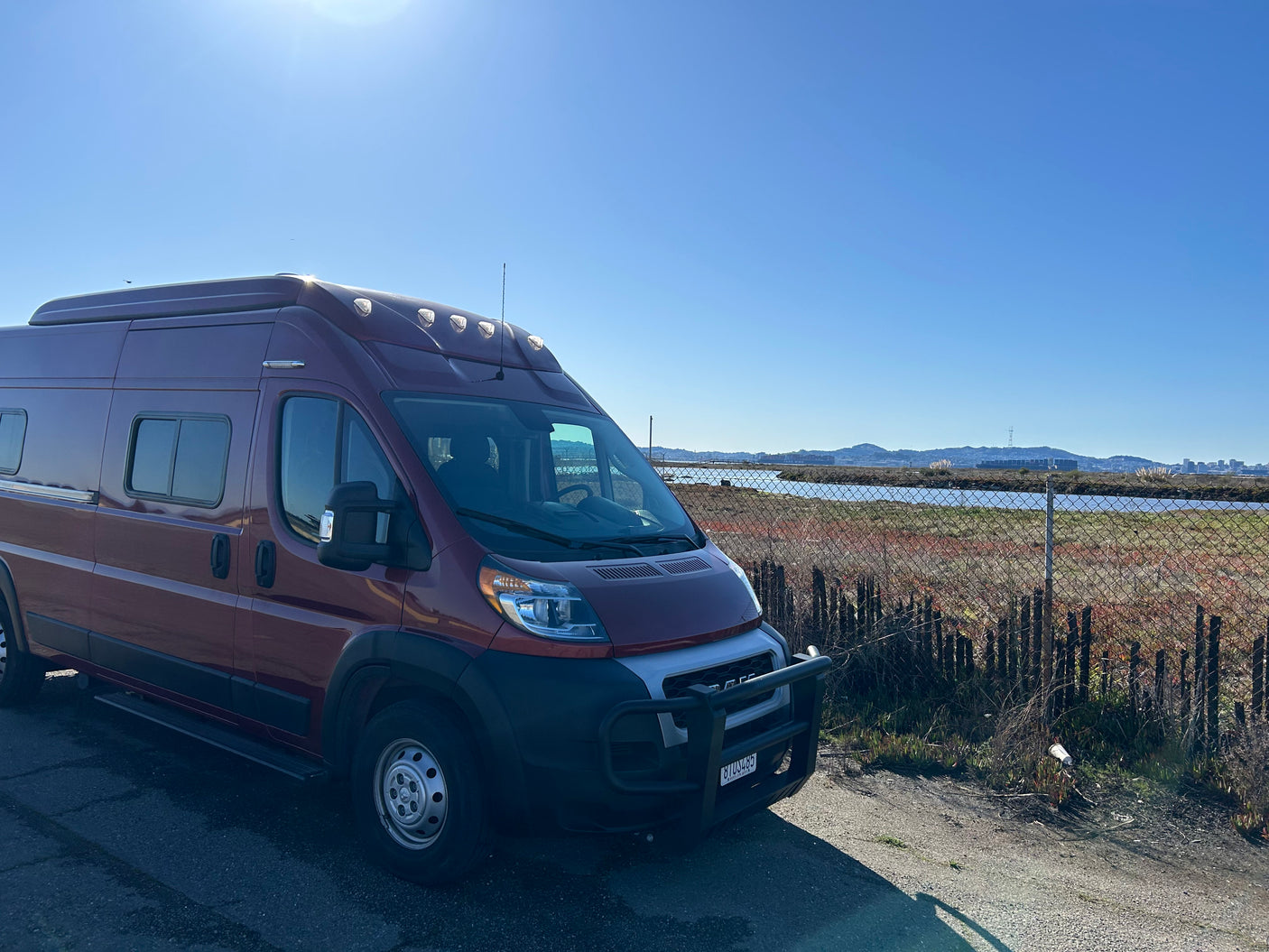 This reddish camper van is parked by water with distant hills & a city skyline, perfect for an RV rental.
