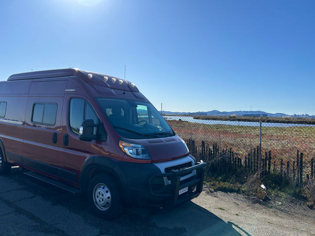 This reddish camper van is parked by water with distant hills & a city skyline, perfect for an RV rental.