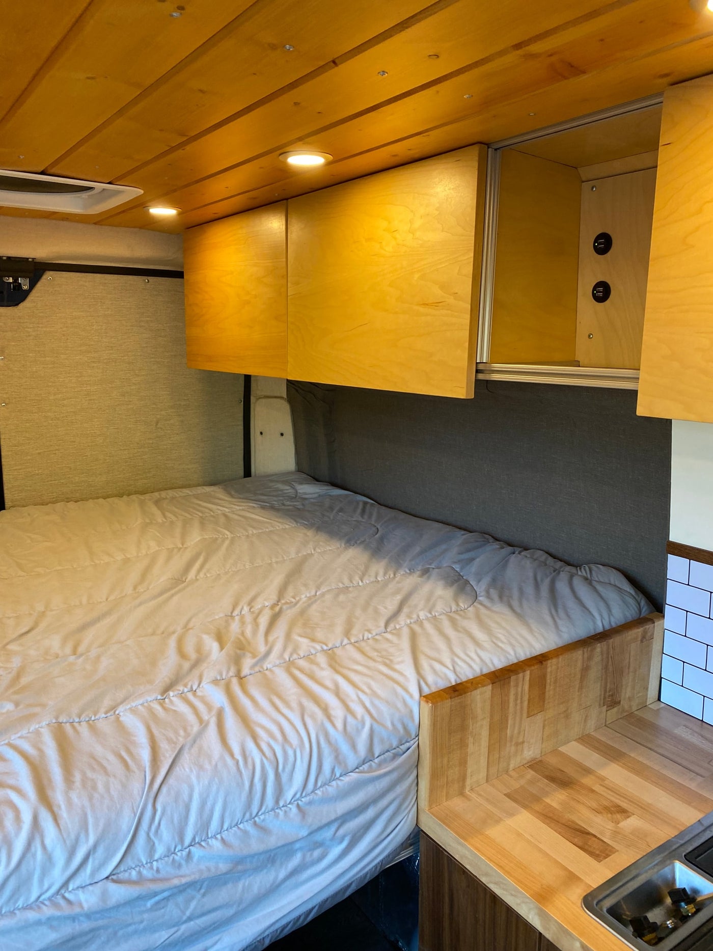 RV rental interior: cozy bed, wood ceiling, overhead cabinets with USB charging, and a small kitchenette with a sink. — photo 7