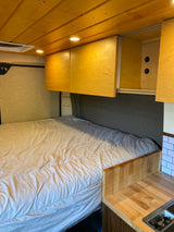 RV rental interior: cozy bed, wood ceiling, overhead cabinets with USB charging, and a small kitchenette with a sink.